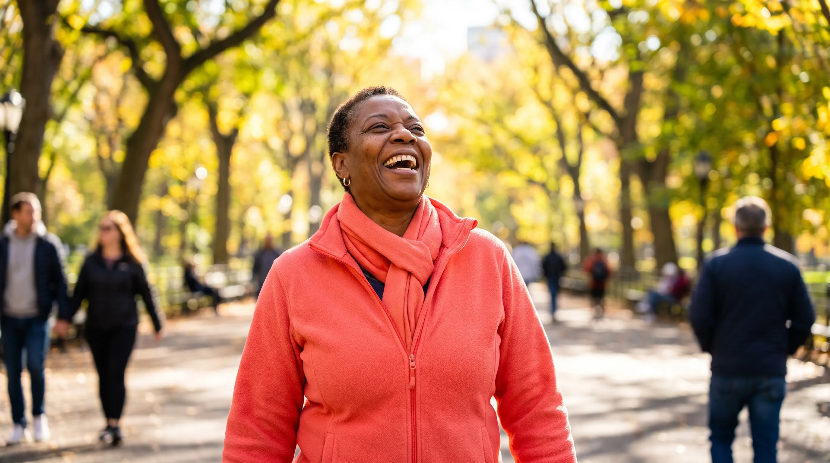 A vital woman in her late fifties laughing on a bright morning walk through a sunlit park, energized and healthy
