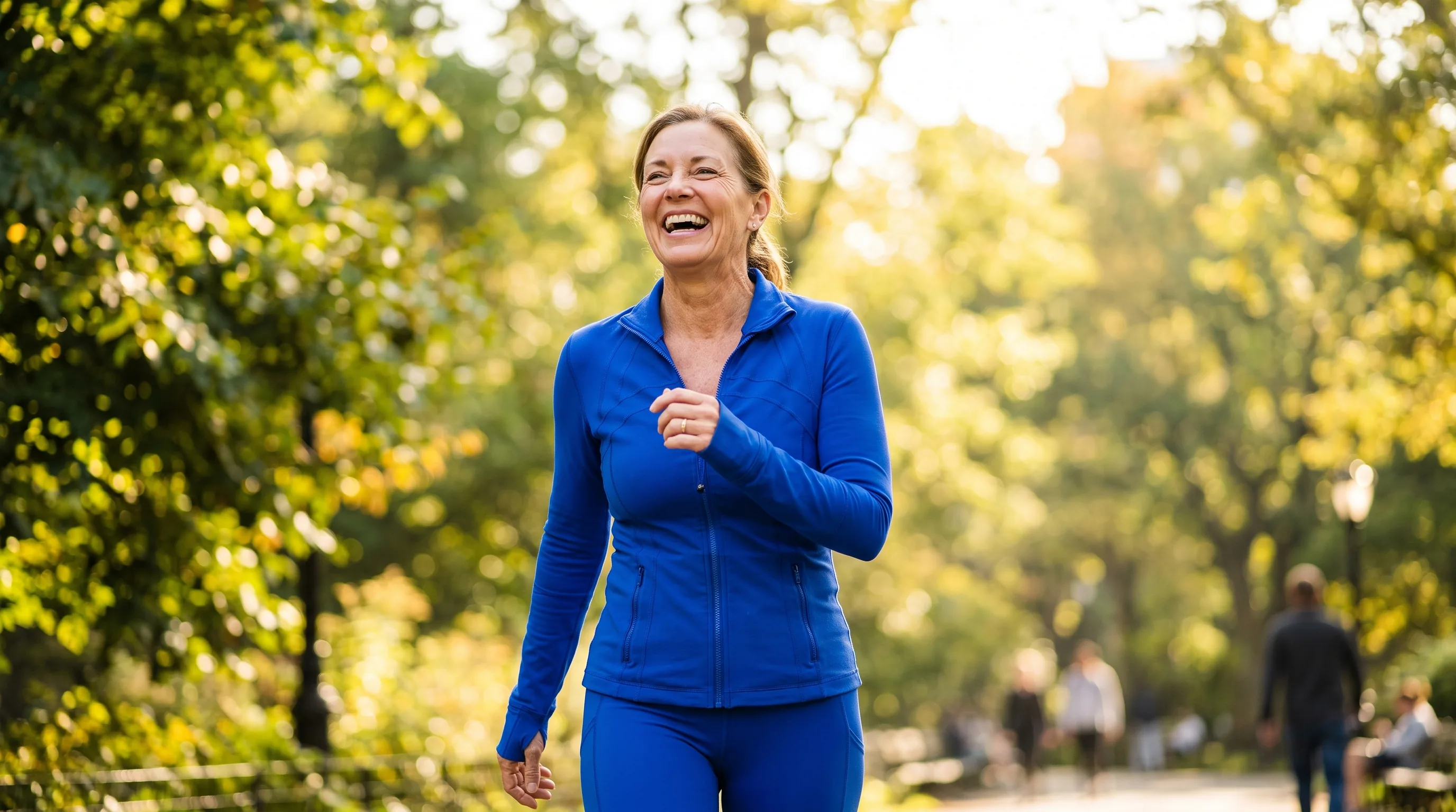 A vital woman in her late fifties laughing on a bright morning walk through a sunlit park, energized and healthy